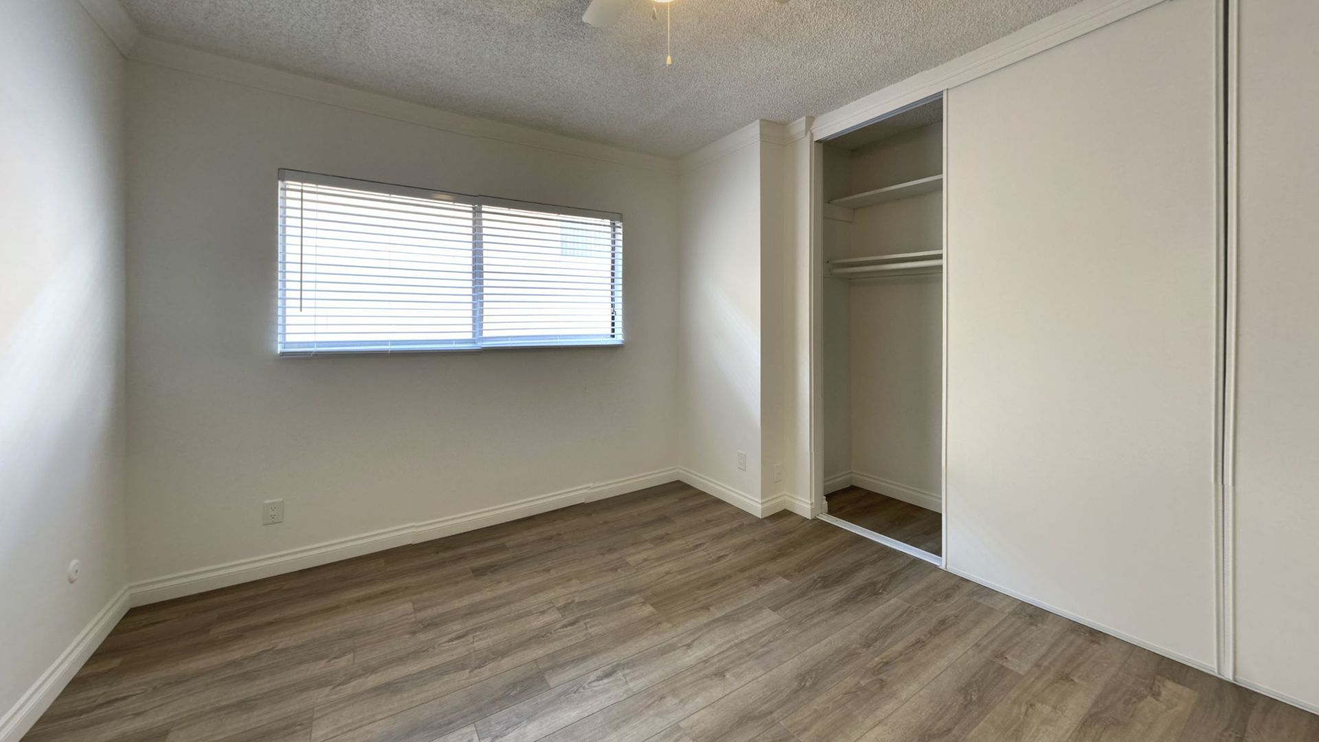 Empty bedroom with wood-look floor, window with blinds, and closet with sliding doors.