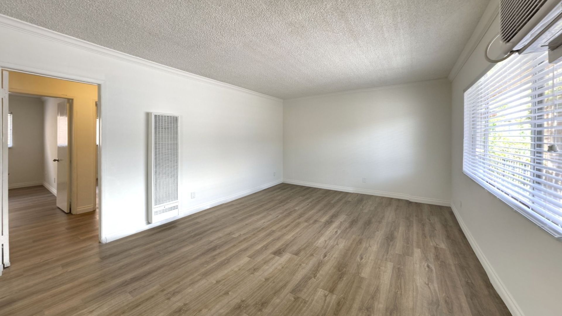 Empty apartment room with wood-look flooring, white walls, and a window with blinds.
