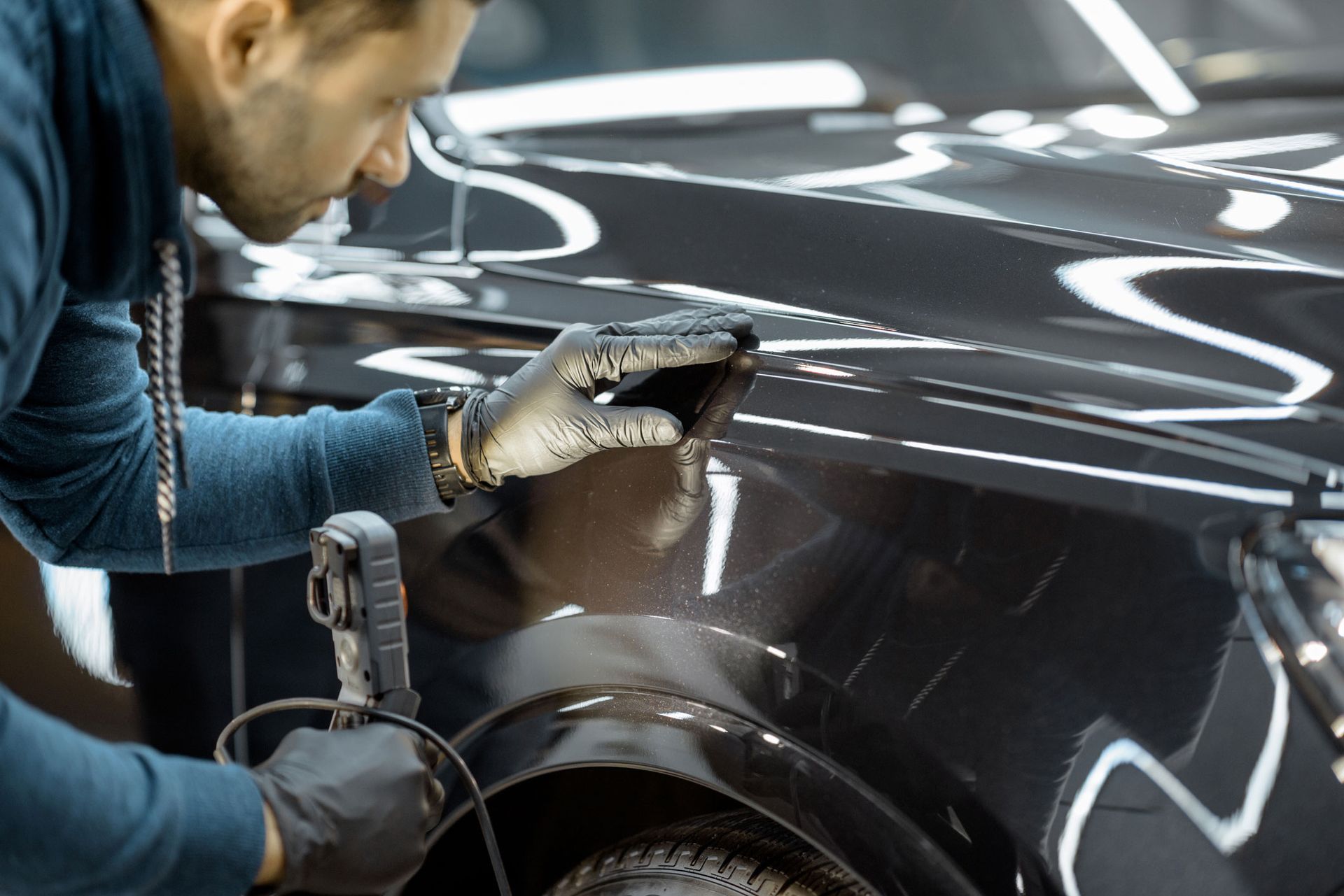 Man in gloves inspecting car paint with a tool, inside a well-lit workshop.