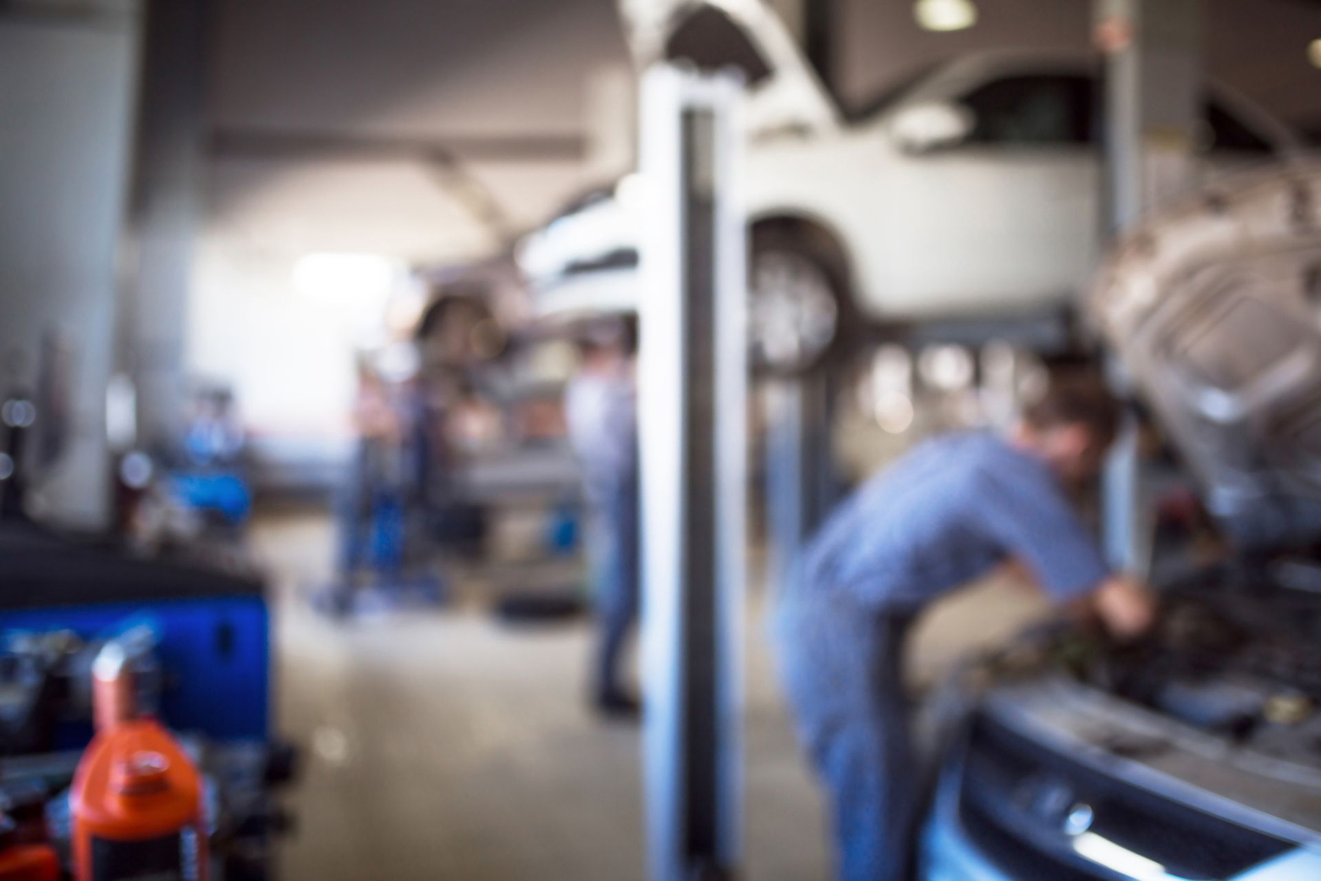 Customer handing car keys to an auto mechanic beside an open vehicle Hood Blurred auto shop with a mechanic working on a car and a red tool in the foreground