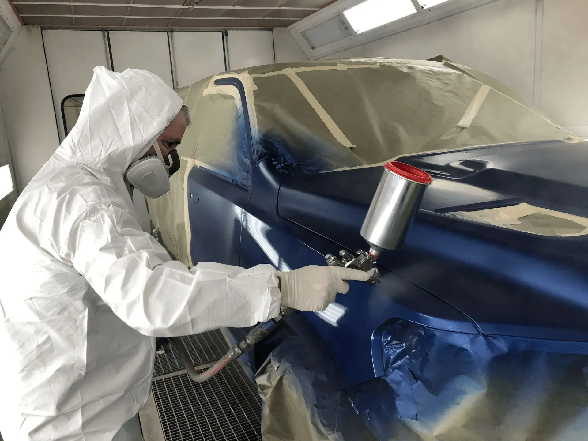 Person in protective suit sprays blue paint on a car in a paint booth.