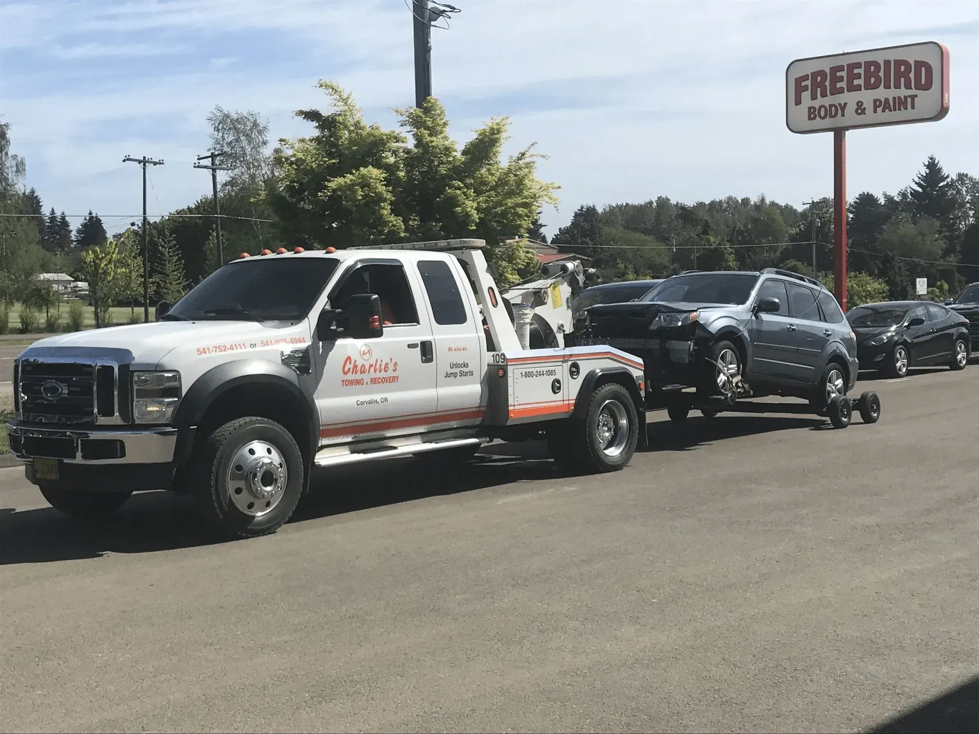 Tow truck pulling a gray SUV on a sunny day, parked in front of a body shop.