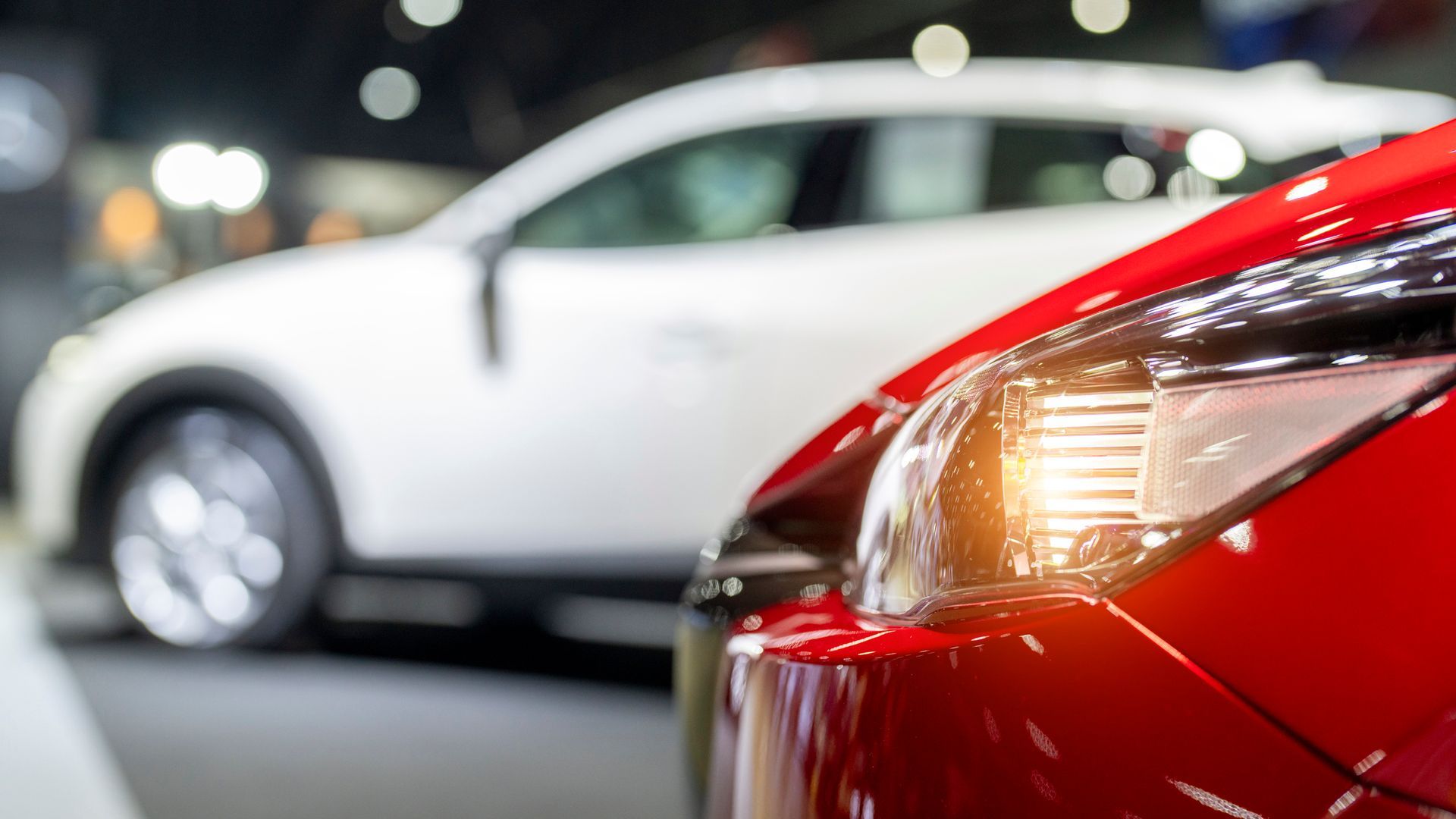 Red car in focus, white car blurred in the background. Cars displayed in a showroom.