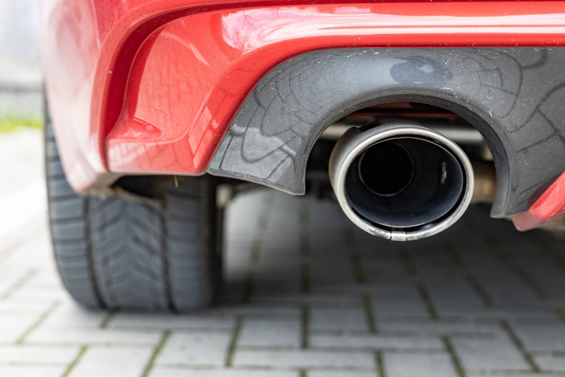 Close-up of a red car's exhaust pipe, black bumper detail, and tire on a brick surface.