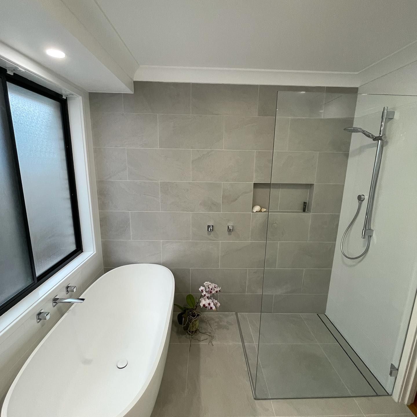 Modern bathroom renovation with freestanding oval bathtub, chrome wall-mounted tapware, frameless glass shower screen, recessed shower niche, large format grey stone-look wall and floor tiles, linear drain, and frosted black-framed window