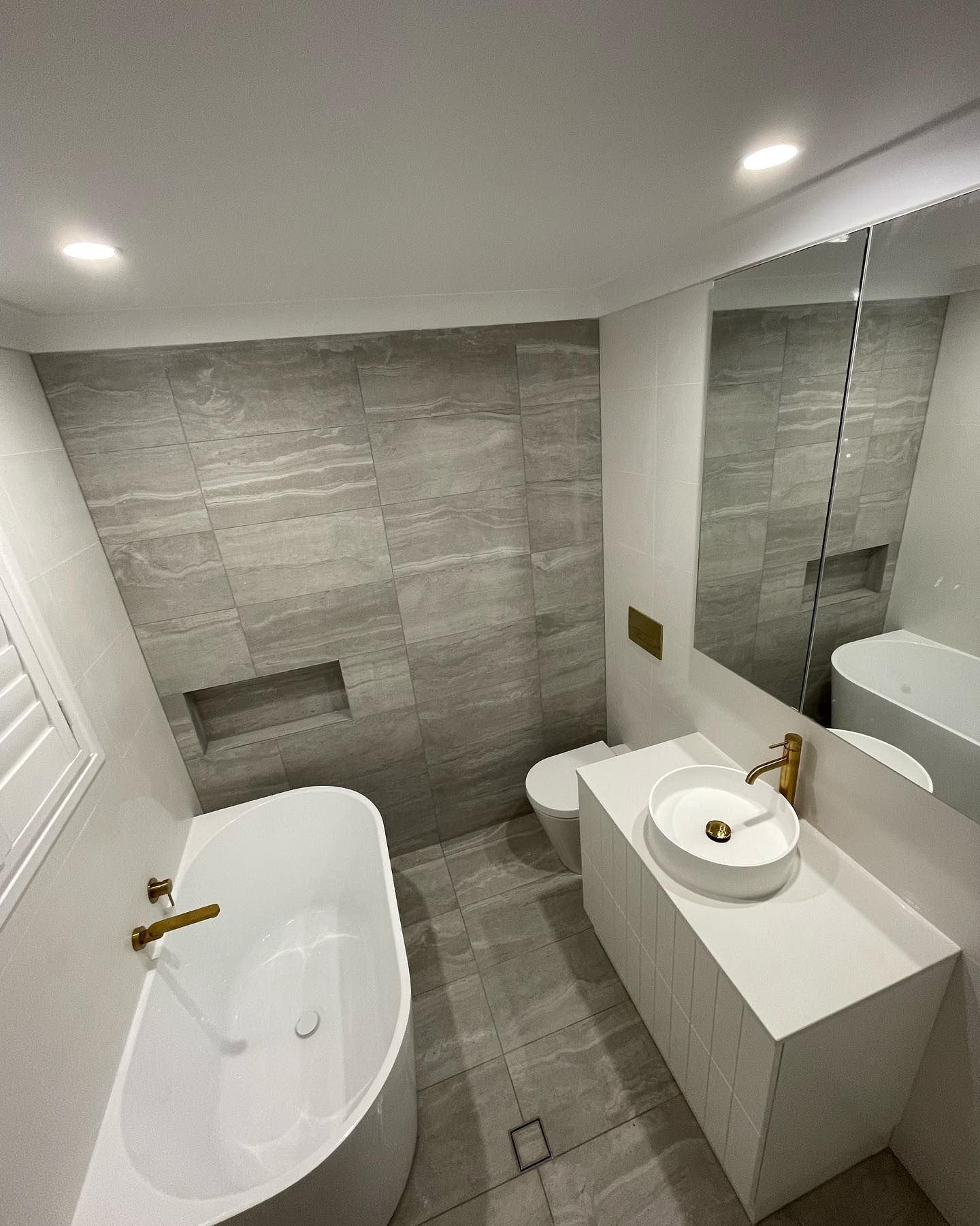 Luxury bathroom renovation with freestanding bathtub, brushed gold tapware, white floating vanity with round vessel sink, large format grey marble-look feature tiles, recessed shower niche, and full-length mirror