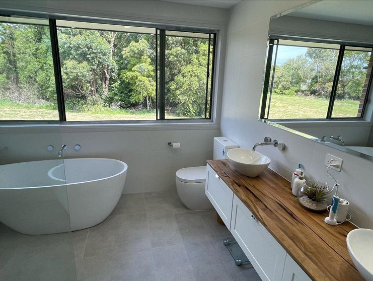Bright modern bathroom renovation with freestanding oval bathtub, live edge timber benchtop on white floating vanity, round vessel sink, wall-mounted chrome tapware, wall-hung toilet, and panoramic black-framed windows with rural garden views