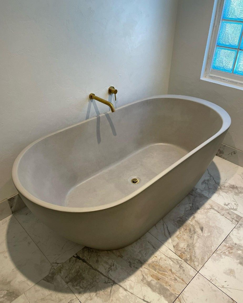 Luxury bathroom featuring a concrete or stone resin freestanding oval bathtub with brushed gold wall-mounted bath mixer and waste, large format marble floor tiles, textured plaster walls, and decorative stained glass window