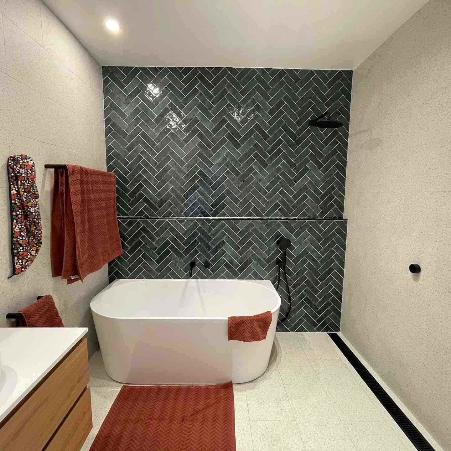 Bold bathroom renovation featuring a back-to-wall freestanding bathtub, dark green gloss herringbone feature wall tiles, terrazzo-look floor tiles, matte black tapware and shower head, timber floating vanity, and linear drain with black trim
