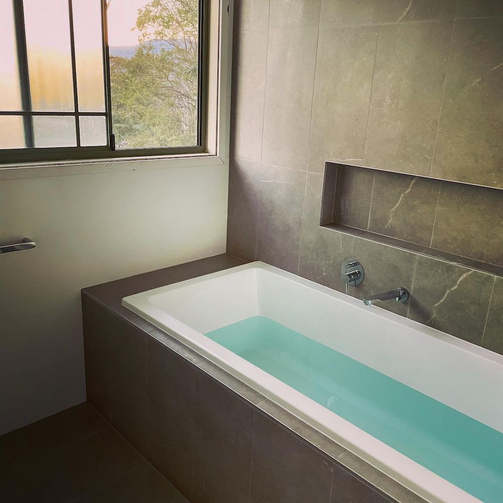 Luxury bathroom renovation featuring a tiled inset bathtub filled with water, chrome wall-mounted mixer tap, recessed bath niche, large format dark grey stone-look marble feature tiles, and black-framed window with mountain and tree views