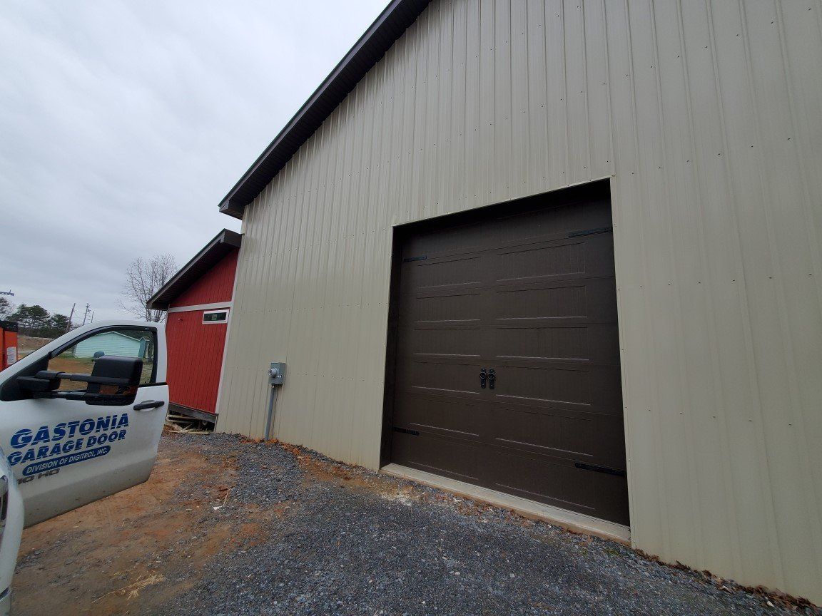 Industrial building with beige siding, a large brown garage door, and a red structure beside a gravel lot.