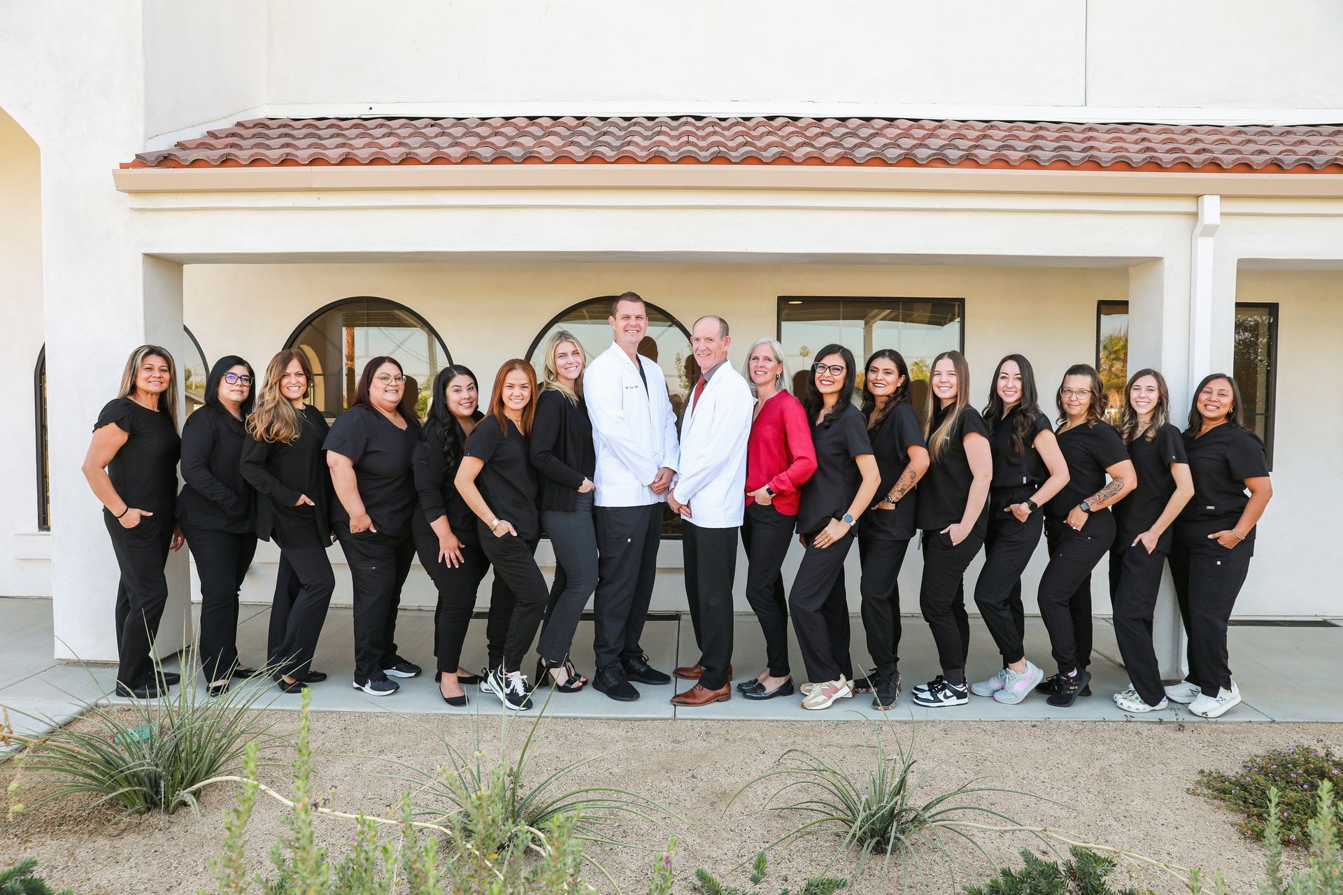 Group of people posing outdoors in front of a modern building, some in black outfits, one in a white coat.