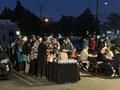 Crowd gathered outdoors at dusk around a table with a black cloth, with string lights in the background