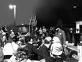 Crowd gathered outdoors at night under streetlights, seated and standing near a waterfront or open plaza.