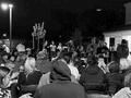 Crowd seated outdoors at night, facing a speaker near a lit stage or platform