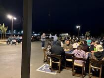 Crowd seated outdoors at night under streetlights, gathered around tables in a parking lot.