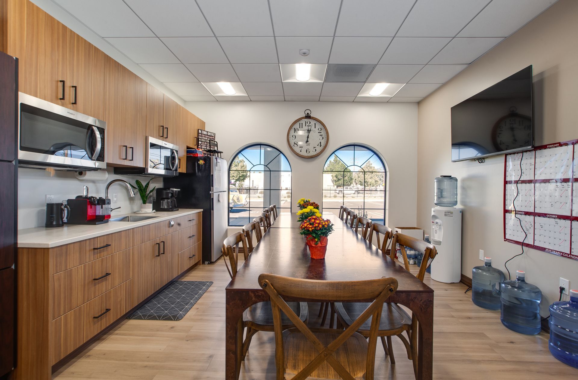 Community kitchen with long wooden tables, cabinets, water dispenser, and arched windows