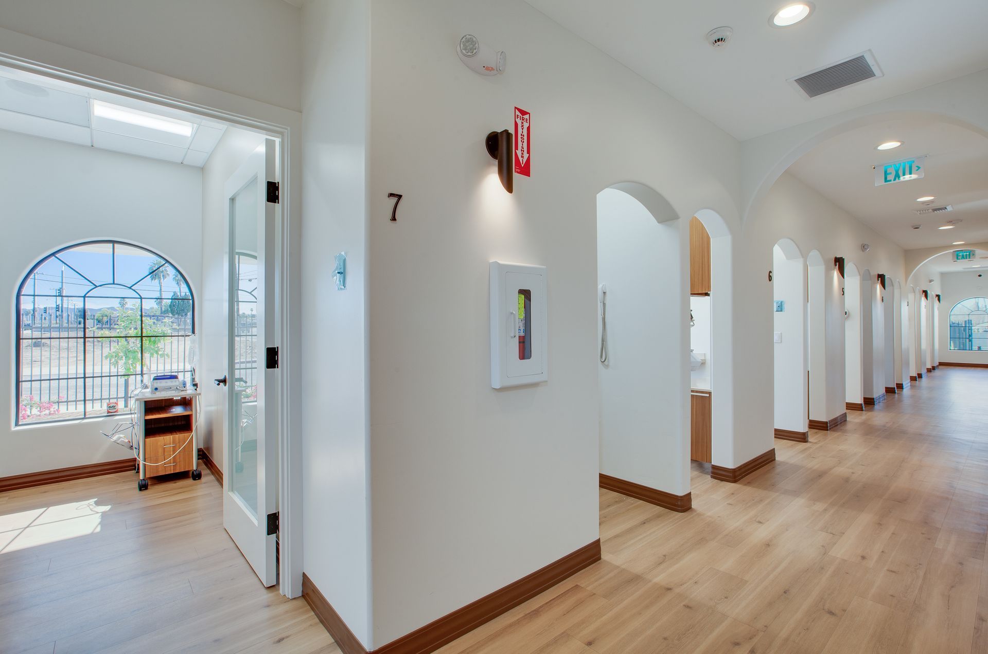 Bright hallway with white walls, wood floors, arched doorways, and a sunlit room at the end.