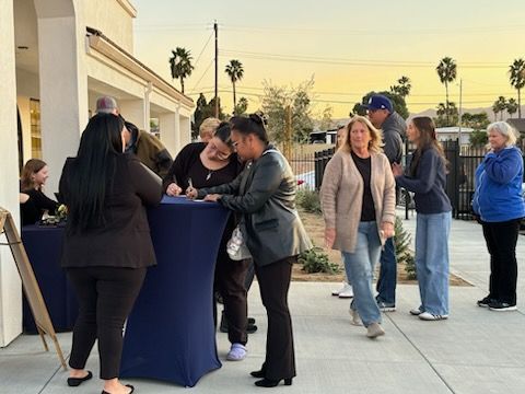 People gathered around a tall blue table outdoors at dusk near a building and palm trees.