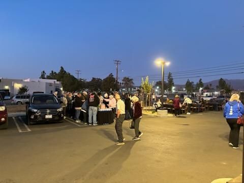 People gathered in a parking lot at dusk, with cars parked and streetlights on.