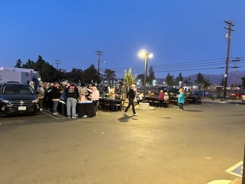 Crowd gathered around parked cars in a lit parking lot at dusk, with people socializing and walking.