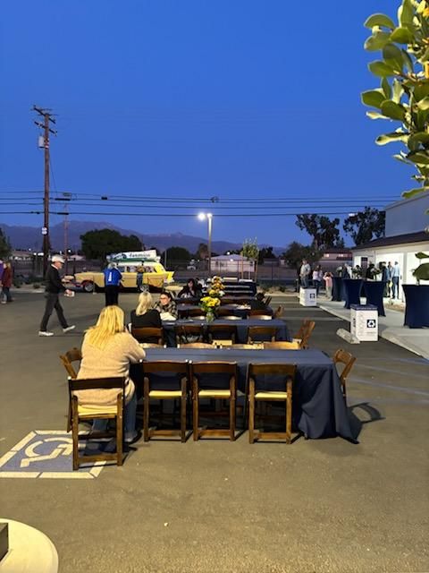 Outdoor dining area at dusk with long tables, benches, and people seated under a blue evening sky