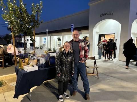 Two people posing outside a lit storefront at night, with a table of items and shoppers in the background