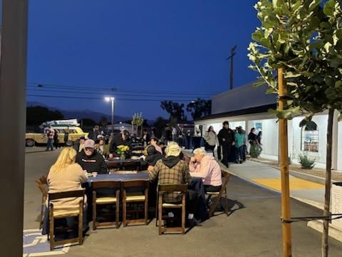 People seated around outdoor tables at dusk beside a lit building and parked cars