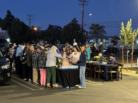 Crowd gathered around a table outdoors at dusk, with people serving food under streetlights.