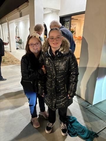 Two children posing indoors by a glass window, one in a black coat and one in a dark hoodie.