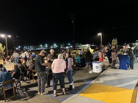 Crowd gathered outdoors at night near tables and tents, with bright lights and a yellow-striped walkway.