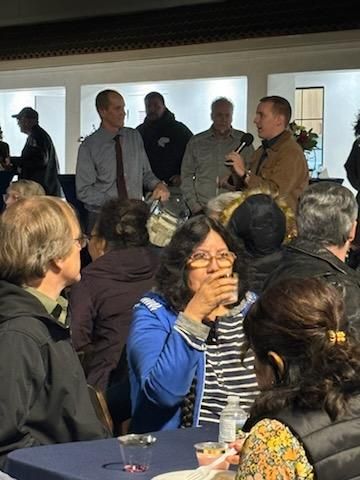 People gathered indoors while a man speaks into a microphone near the front.