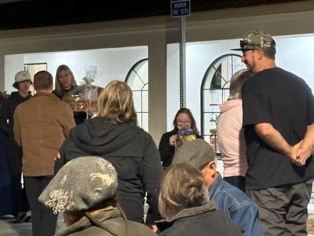 People gathered indoors near a display table and arched window, with one person speaking to the group.