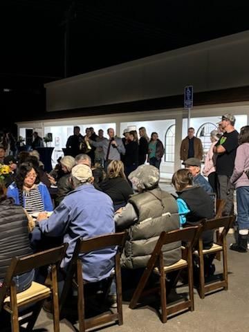 People seated outdoors at night, facing a small crowd gathered under a lit storefront.