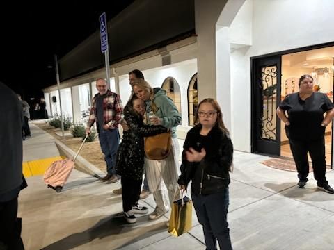 People standing outside a lit storefront at night; a woman in glasses stands in the foreground holding a yellow bag.