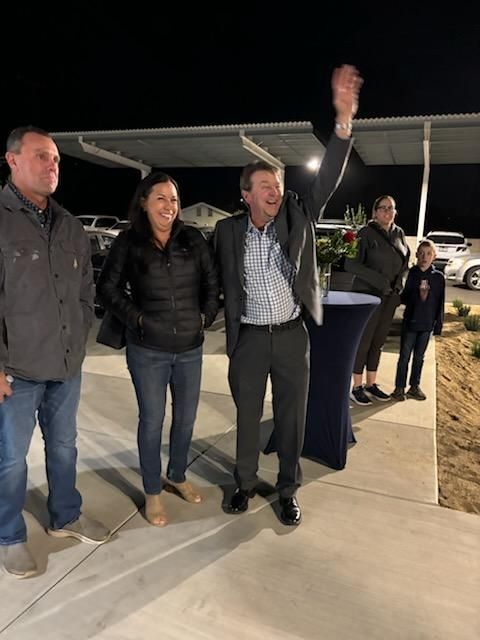 Four people stand outdoors at night, one waving beside a high-top table and parked cars.