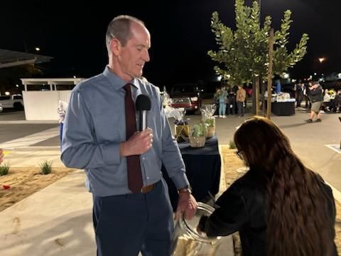 Man speaking into a microphone while kneeling woman reaches toward him at an outdoor nighttime event