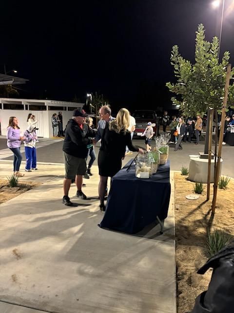 People gathered around a table at a nighttime outdoor event, with a tree and buildings in the background.