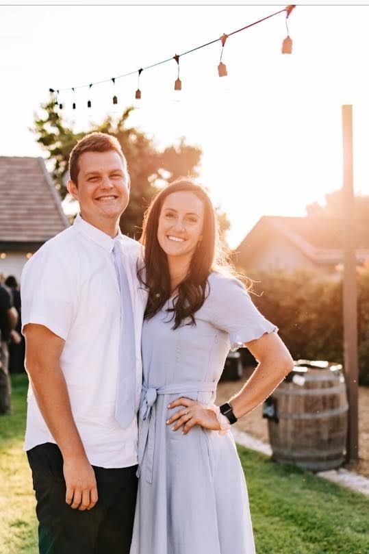 Two people standing outdoors at sunset, smiling in white and light blue outfits.