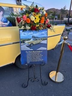 Yellow vintage car with floral wreath and commemorative sign on an easel at an outdoor event