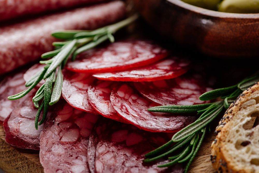 Salame affettato su un tagliere di legno con rametti di rosmarino fresco, pane e una ciotolina di olive.