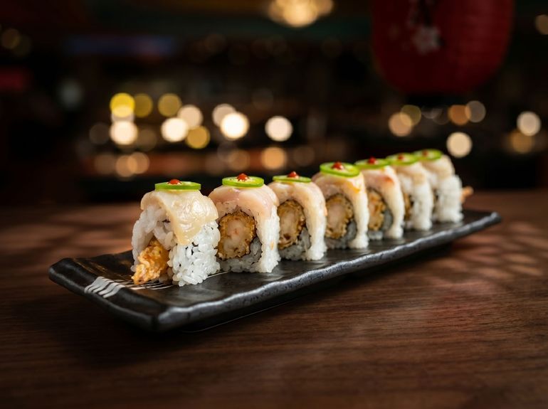 Sushi roll topped with creamy sauce and garnish on a black platter, blurred warm lights in background