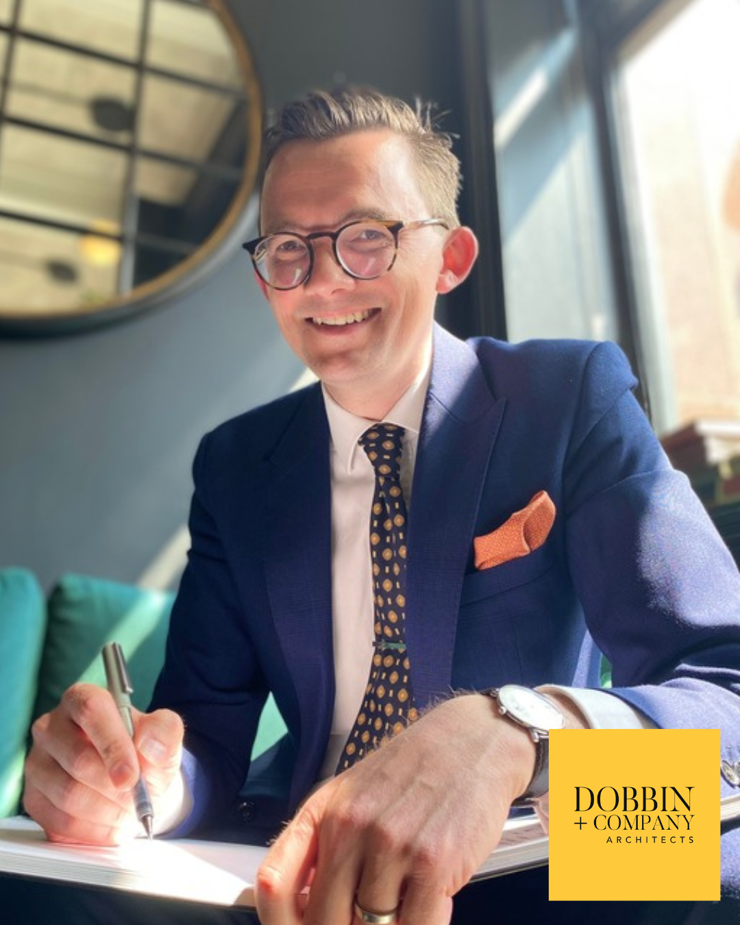 John Dobbin in blue suit writing at a table; smiling with glasses, orange pocket square.