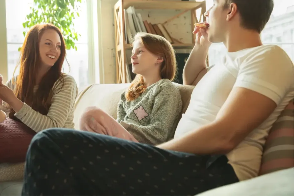 A person sits on a sofa with a parent and child who are eating together in a brightly lit living room.