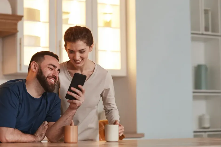 A man and woman lean over a counter together, smiling while looking at a smartphone held by the woman.