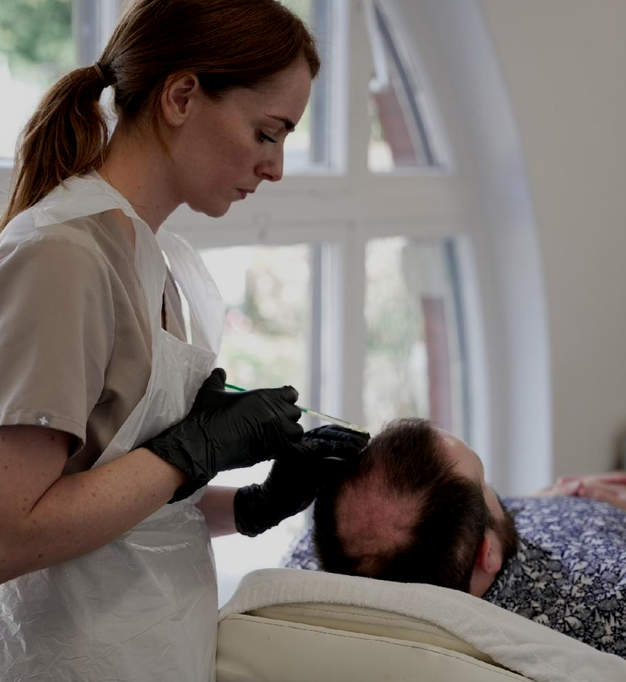 A woman in gloves performs a scalp micropigmentation procedure on a man lying down in a salon.