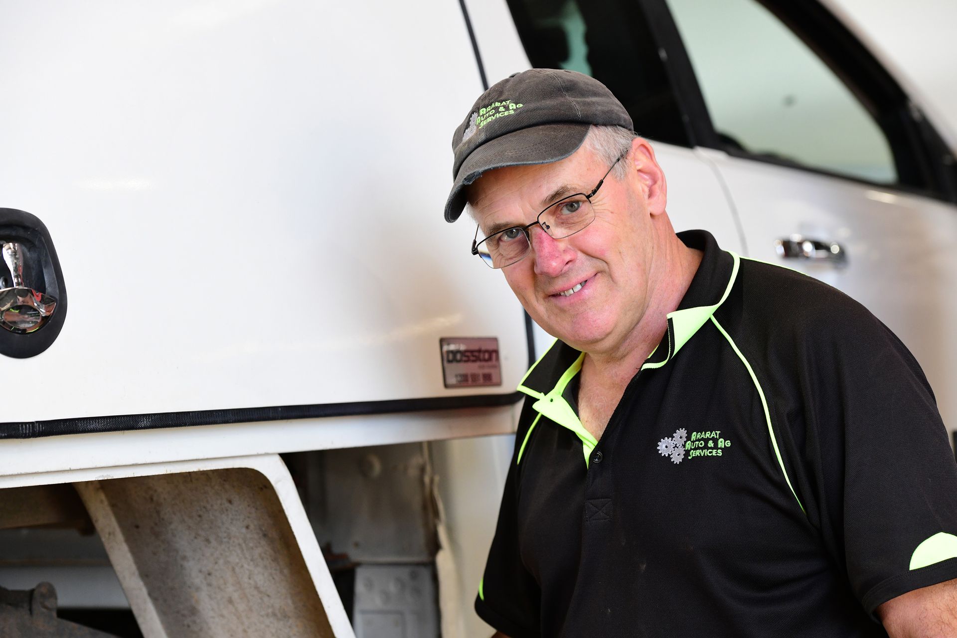 Man in black shirt and cap leaning beside a white truck, smiling at the camera.