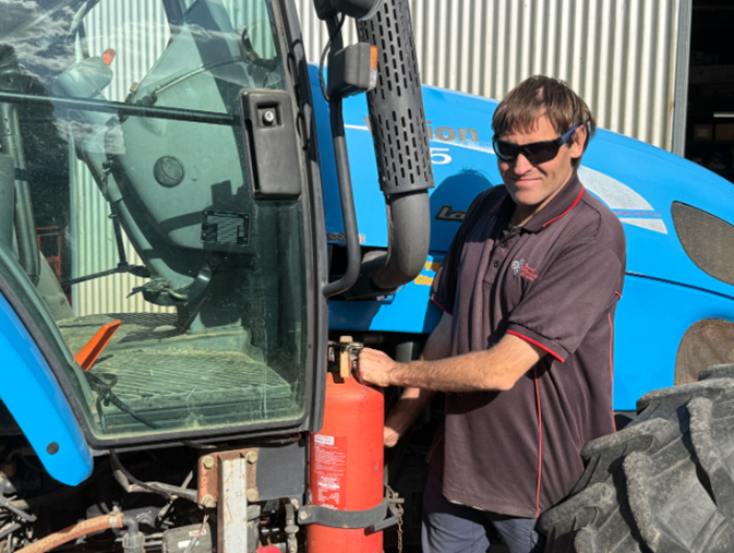 A man in a polo shirt and sunglasses standing next to a blue tractor, placing a red fire extinguisher into a mounting bracket. A man in a polo shirt and sunglasses standing next to a blue tractor, placing a red fire extinguisher into a mounting bracket.
