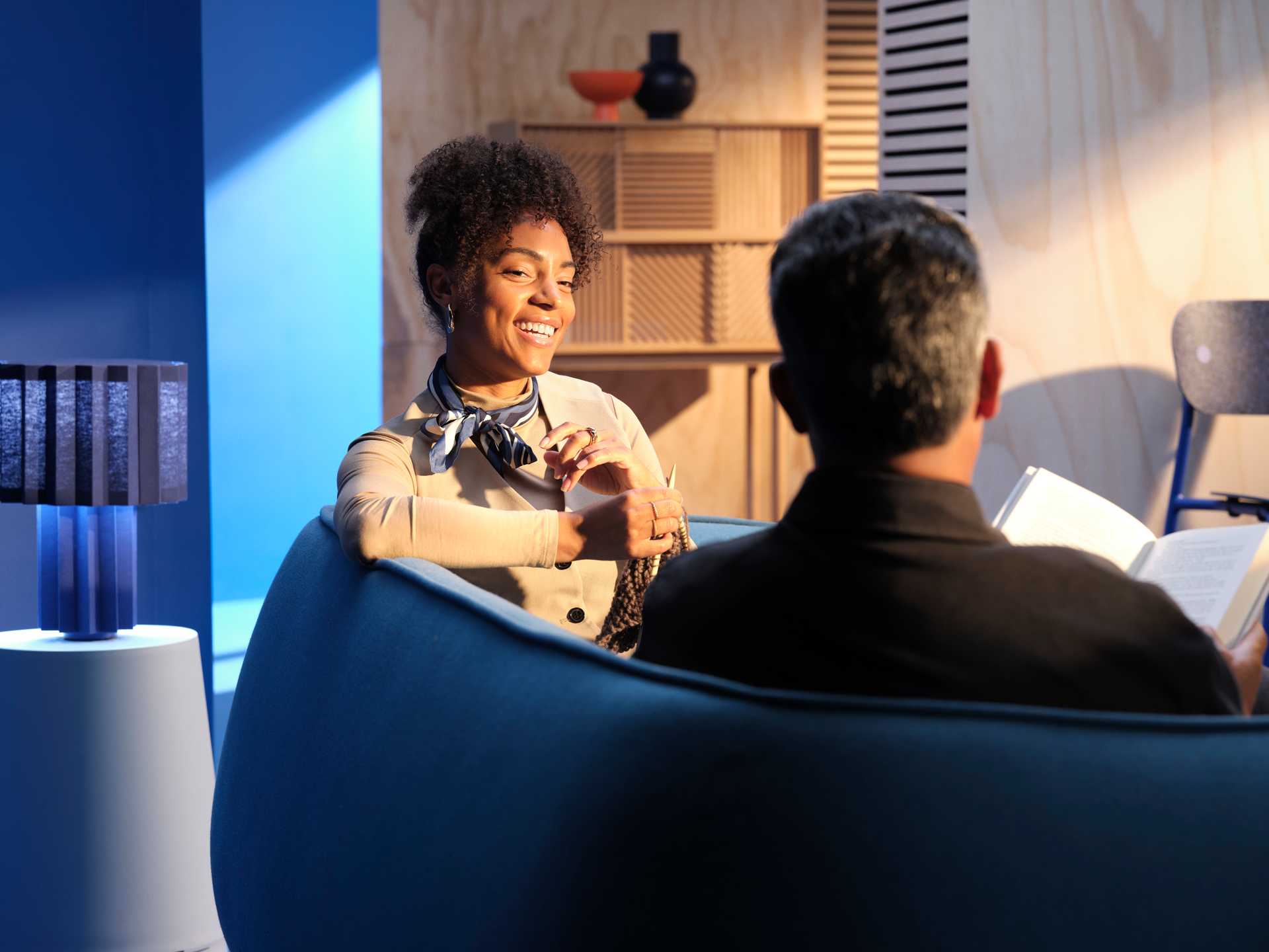 Two people in a cozy meeting room, one speaking from a blue chair with a lamp and notebook nearby.