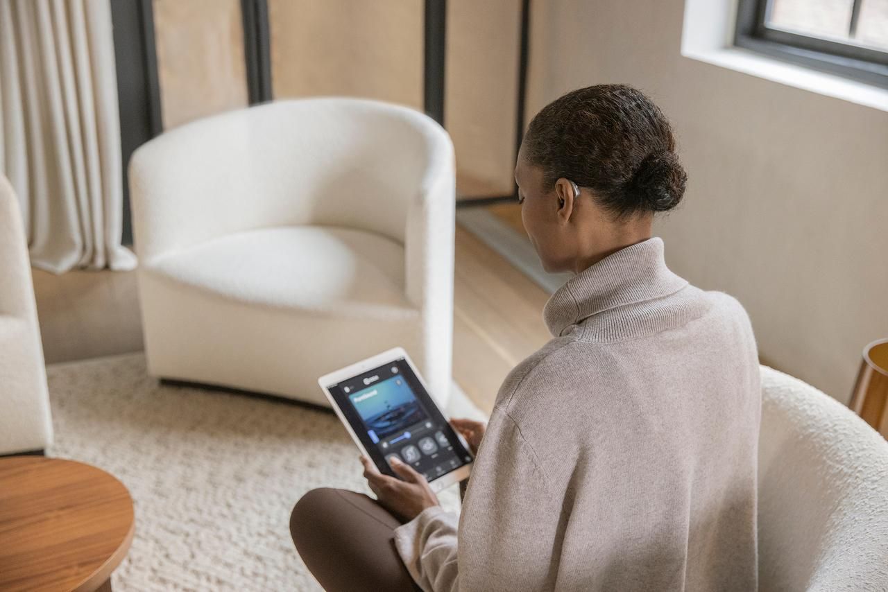 Person seated in a bright room, using a tablet beside a white armchair and window curtains